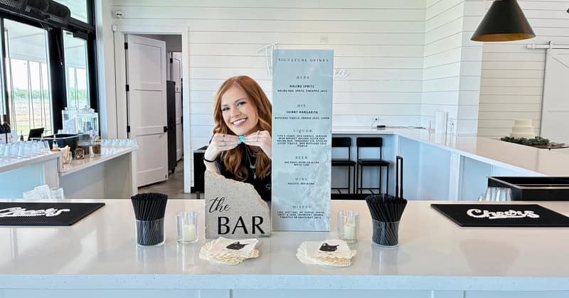 White bar set up indoors with a framed drinks menu, stacked glassware and cocktails poured ready to serve