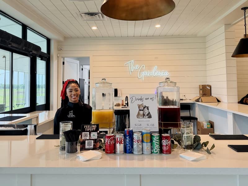 Shaker bartender smiling behind a fully set white bar with drink dispensers, canned sodas and signature cocktail signage