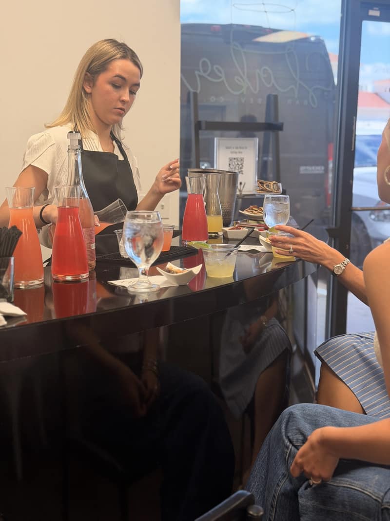 Shaker bartender in a black apron making drinks behind a bar while seated guests wait with their glasses
