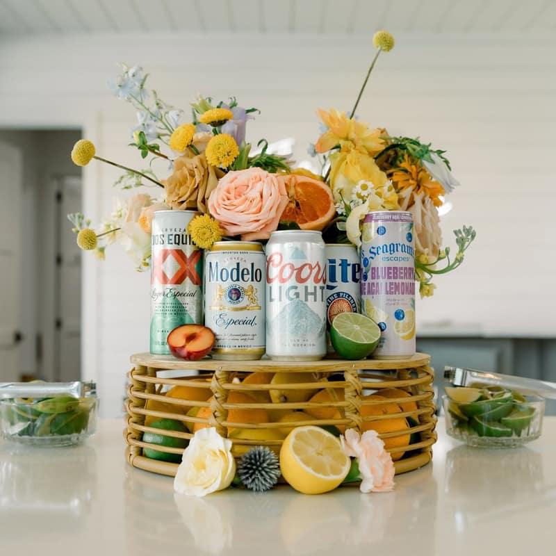 Cans of beer and hard lemonade on a rattan riser surrounded by yellow florals, sliced citrus and bowls of limes
