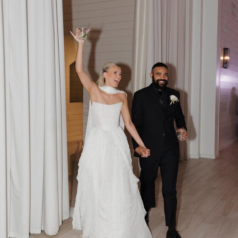 Bride holding a coupe glass aloft as she and the groom walk into their reception hand in hand