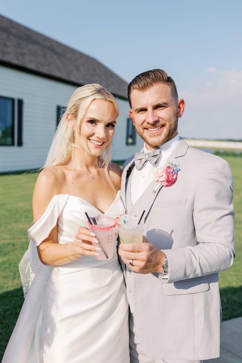 Bride and groom smiling and holding personalized wedding cups in front of a white barn venue