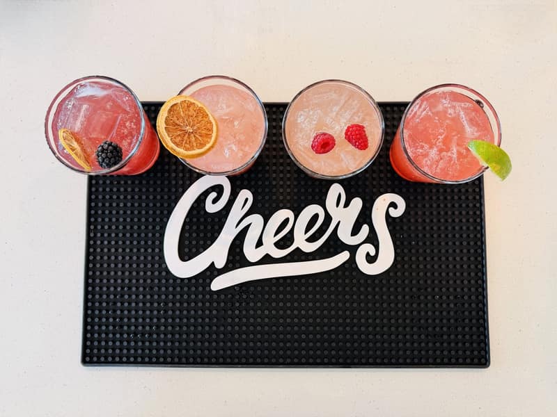 Four garnished cocktails lined up on a black bar mat above a Cheers sign, photographed from above