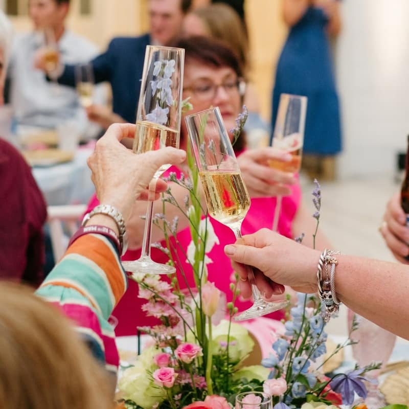 Party guests reaching in to clink champagne flutes above a table of pink and blue florals