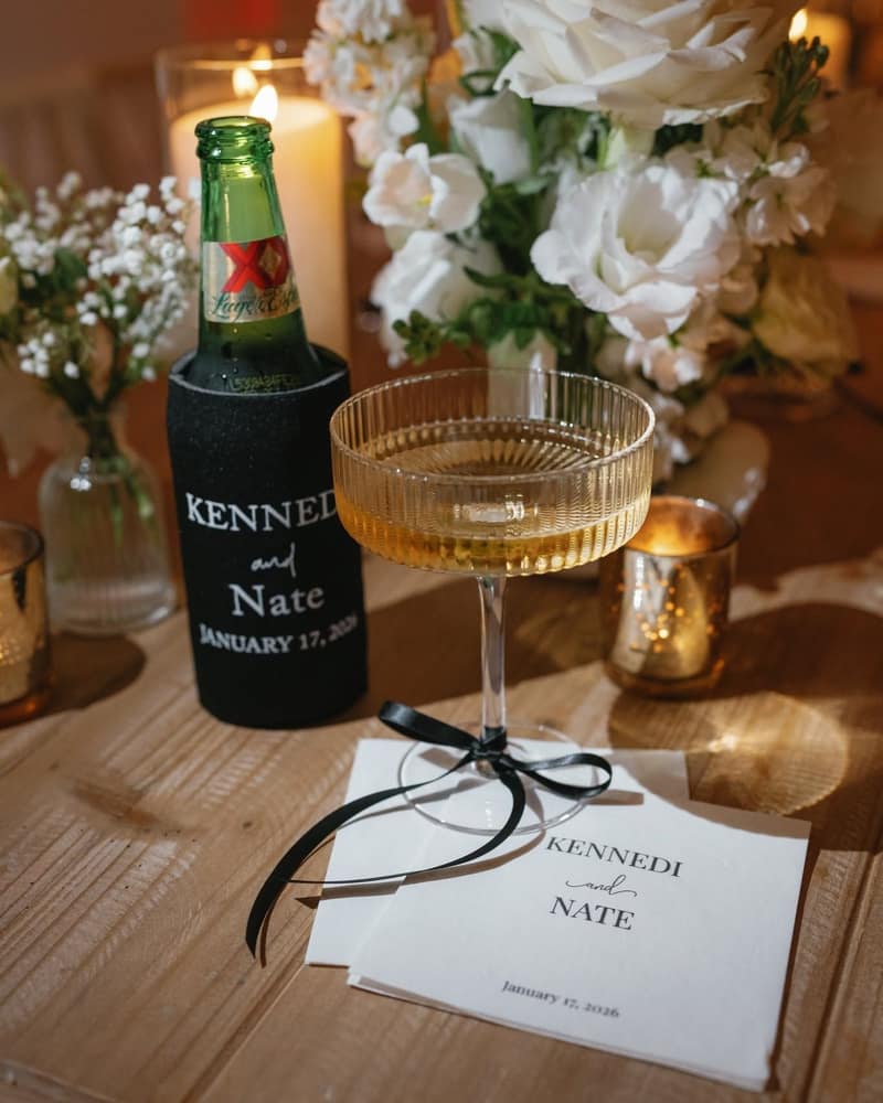 Personalized koozie on a beer bottle beside a fluted coupe of champagne and printed napkins on a wooden reception table