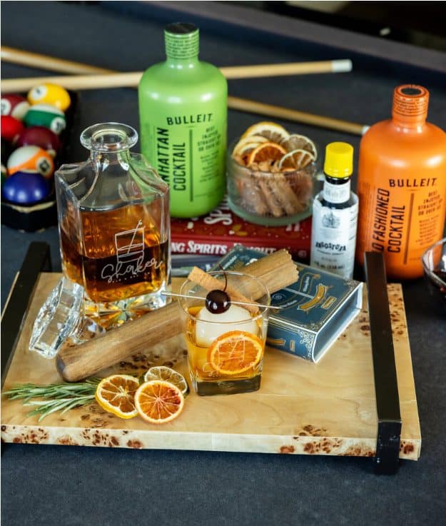 Etched Shaker decanter and a smoked old fashioned on a wood tray with Bulleit bottles and bitters, set up beside a game room pool table