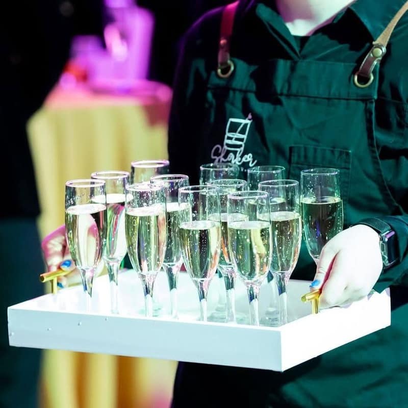 Bartender in a Shaker apron carrying a tray of filled champagne flutes out to guests