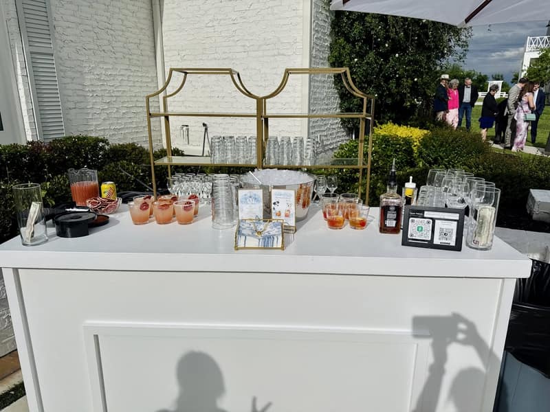 White portable bar set up outdoors with glassware on a gold shelf and cocktails poured, guests gathered behind